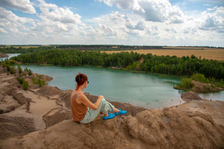 A woman with red hair and dark glasses sits on a high mountain above a bright blue lake .の写真素材