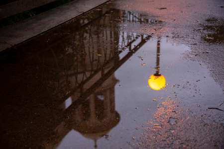A bright yellow lantern, part of the temple and openwork railing are reflected in a rain puddle on the asphalt in the evening.の写真素材
