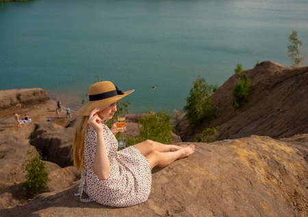 A blonde girl in a straw hat and summer dress is sitting with a glass of champagne on a high hill above the lake.の写真素材