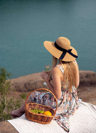 A girl with long blond hair in a hat and a smart sundress sits with a glass of champagne on a hill with her back to the camera.There is a fruit basket next to it.の写真素材