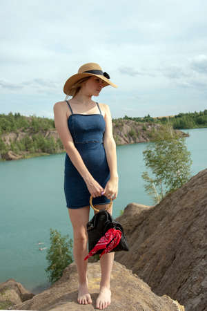 A young girl with blonde hair, wearing a denim sundress stands on a high hill above a blue lake.の写真素材