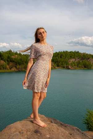 A blonde girl in a short summer dress stands on a high Bank above a blue lake.の写真素材