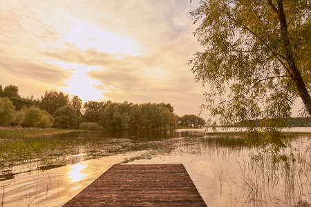 Early autumn evening landscape. The sun is reflected in the surface of the lake, surrounded by trees. In the foreground are wooden bridges.の写真素材