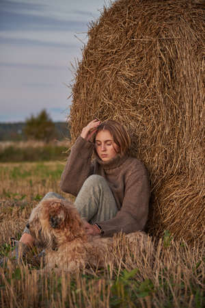 A beautiful sad blonde girl and a dog are sitting in a field.の写真素材