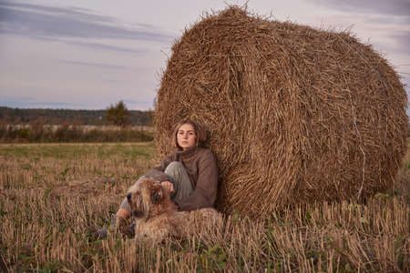 A beautiful sad blonde girl and a dog are sitting in a field, leaning against a haystack.の写真素材