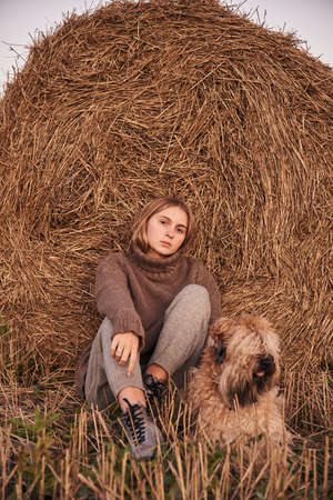 A blonde girl in a warm knitted sweater and a dog sit next to a haystack in the evening.の写真素材