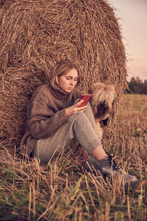 A blonde girl in a warm knitted sweater with the phone in hand and a dog sit next to a haystack in the evening.の写真素材