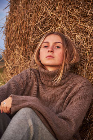 Tinted portrait of a charming young girl in a warm sweater, sitting by a haystack. Stylish girl relaxing on hay bale in autumn field in sunset.の写真素材