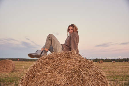 Tinted portrait of a charming young girl in a warm sweater, sitting on a haystack .. Stylish girl relaxing on hay bale in autumn field in sunset.の写真素材