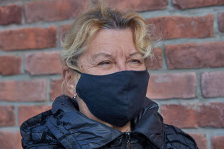 Portrait of an elderly woman in a black medical mask against a brick wall.の写真素材