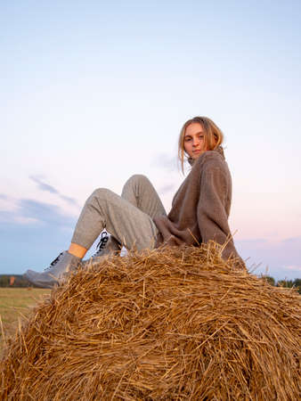 portrait of a charming young girl in a warm sweater, sitting on a haystack .. Stylish girl relaxing on hay bale in autumn field in sunset.の写真素材