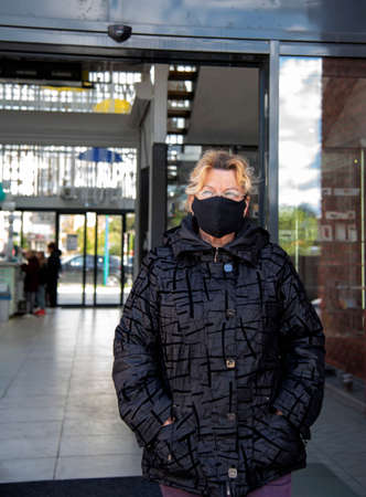 An elderly woman in a black jacket and a black medical mask walks out of the door of a shopping center on a Sunny autumn day.の写真素材
