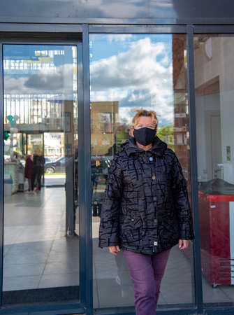 An elderly woman in a black jacket and a black medical mask walks out of the door of a shopping center on a Sunny autumn day.の写真素材