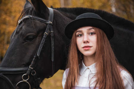 Red-haired girl in a black hat with a Chestnut horse in the autumn forest.の写真素材