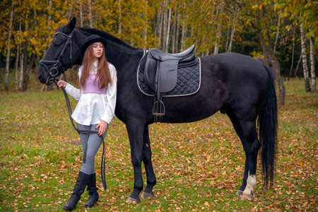 Red-haired girl in a black hat with a Chestnut horse in the autumn forest.の写真素材
