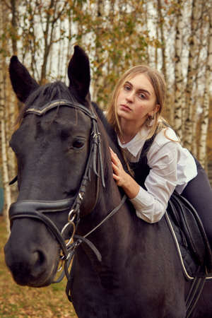 Charming blonde in a white shirt riding a horse gently hugs her neck against the background of the autumn forest.の写真素材