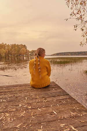 A teenage girl sits on the edge of a wooden bridge over the lake with her back to the camera against the sunset.の写真素材