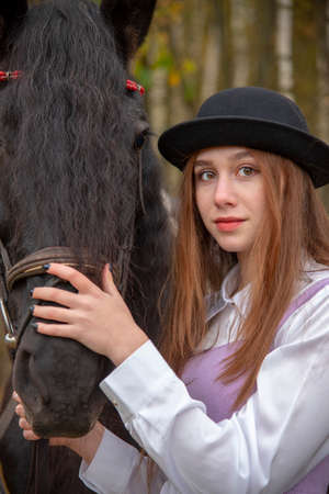 Red-haired girl in a black hat with a Chestnut horse in the autumn forest.の写真素材
