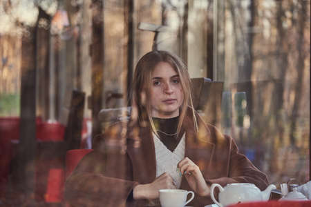 Slightly blurry portrait of a charming girl, made through the window of a cafe. The window reflects the autumn Park.の写真素材