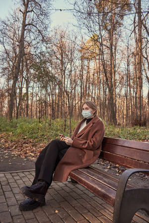 A charming girl in a medical protective mask with a phone in her hands is sitting on a bench in an autumn Parkの写真素材