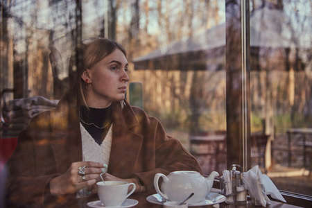 Slightly blurry portrait of a charming girl drinking tea, made through the window of a cafe. The window reflects the autumn Park.の写真素材