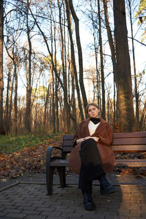 A charming girl with blonde hair, wearing a brown wool coat, is sitting on a wooden bench in an alley in an autumn Park. Autumn photo sessions in nature.の写真素材