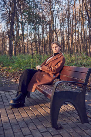 A charming girl with blonde hair, wearing a brown wool coat, is sitting on a wooden bench in an alley in an autumn Park. Autumn photo sessions in nature.の写真素材