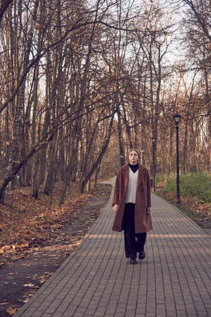 A blonde charming girl in a brown coat walks along an alley in an autumn Park.の写真素材