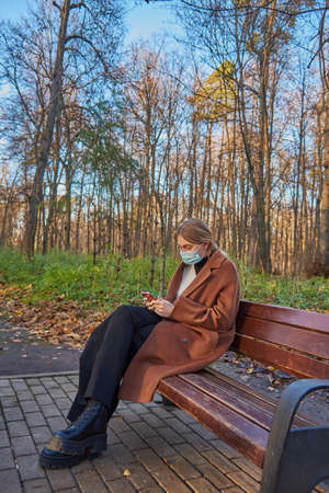 A charming girl in a medical protective mask with a phone in her hands is sitting on a bench in an autumn Parkの写真素材