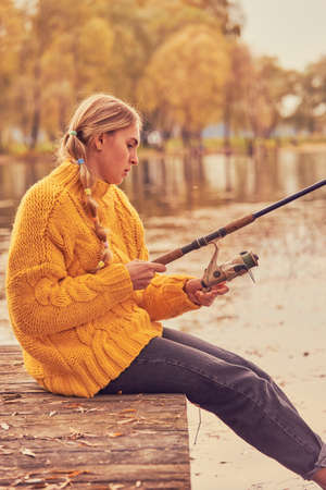 A girl with pigtails in a bright yellow knitted sweater sits on a wooden bridge over the lake and catches fish.の写真素材