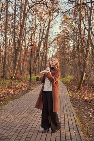 A blonde charming girl in a brown coat walks along an alley in an autumn Park.の写真素材