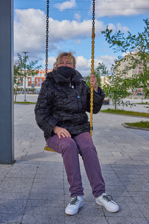 A 68-year-old woman wearing a black medical protective mask sits on a swing in the town squareの写真素材