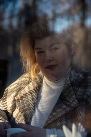Portrait of a plump middle-aged woman with blonde hair, taken through the window of a cafe, which reflects the bare autumn trees.の写真素材