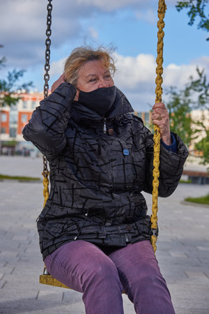 A 68-year-old woman wearing a black medical protective mask sits on a swing in the town squareの写真素材
