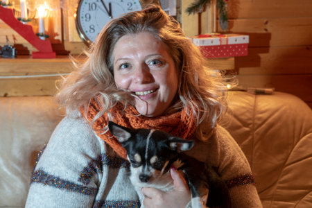 a plump, middle-aged blonde woman sits on a leather sofa with a small dog, a chihuahua, in a room with a christmas decor.の写真素材