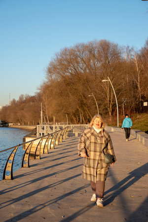 A beautiful plump blonde woman in a plaid coat walks along the embankment on a clear late autumn day.の写真素材