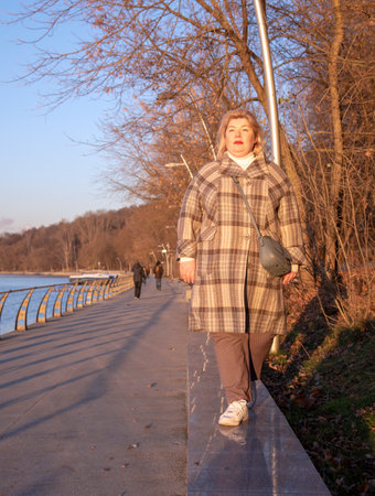 A beautiful plump blonde woman in a plaid coat walks along the embankment on a clear late autumn day.の写真素材