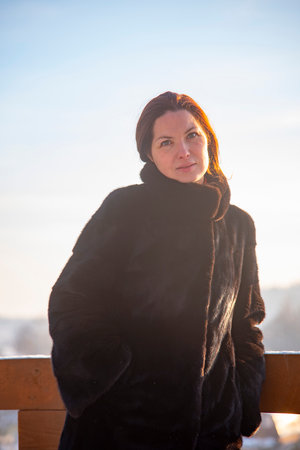A middle-aged woman with red hair , wearing a black fur coat, against the background of a winter landscape on a sunny day. Portrait in a sunny haze.の写真素材