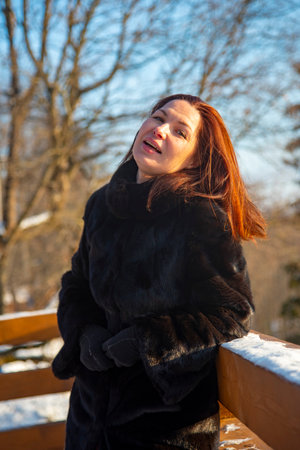 A middle-aged woman with red hair , wearing a black fur coat, against the background of a winter landscape on a sunny day.の写真素材
