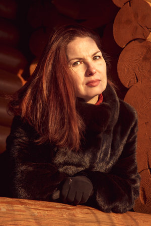 A beautiful 45-year-old woman with red hair poses on the veranda of a log house on a winter day.の写真素材