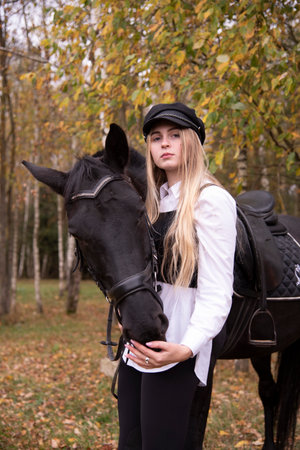 Portrait of a teenage girl with long blonde hair and a black cap, hugging a Chestnut horse against the background of an autumn forest.の写真素材
