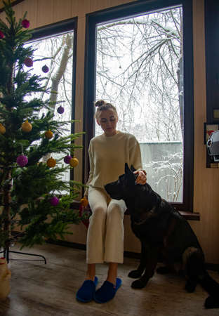 A beautiful blonde girl in a white house suit and a shepherd dog at the window with a view of the snow-covered trees.の写真素材