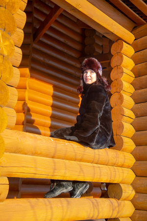 A middle-aged woman in a fur hat looks out of the window of a log house.の写真素材