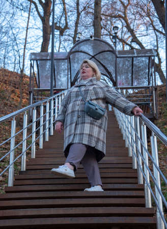 A plump, blonde woman of 45 years in a fashionable plaid coat climbs the stairs in a late-autumn public park.の写真素材