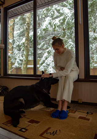 A beautiful blonde girl in a white house suit and a shepherd dog at the window with a view of the snow-covered trees.の写真素材