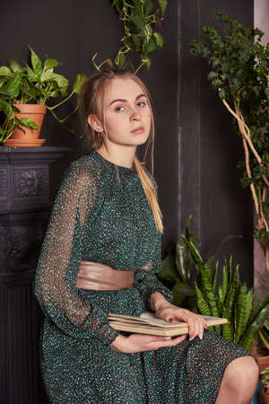 A blonde girl in a chiffon green dress with a book in hand in a cozy room.の写真素材