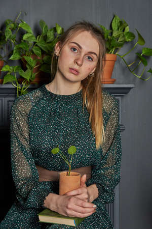 A blonde girl in a chiffon green dress with a flower pot in her hands on the background of a gray wall.の写真素材