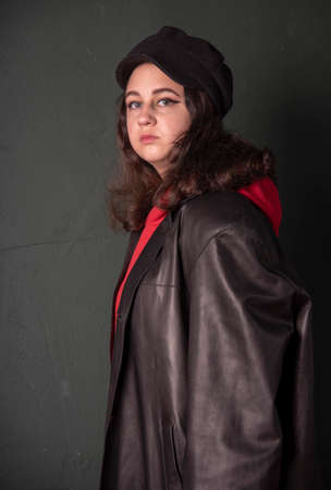A girl in a black hat, wearing a red hoodie and a black leather jacket on the background of a dark wall. Photo shoot in the studio. Youth trend.の写真素材