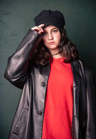 A girl in a black hat, wearing a red hoodie and a black leather jacket on the background of a dark wall. Photo shoot in the studio. Youth trend.の写真素材