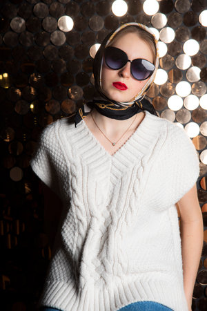 A girl in a white knitted vest, dark glasses and a silk scarf against the background of a golden wall. Fashion trends, photo shoot in the studio.の写真素材